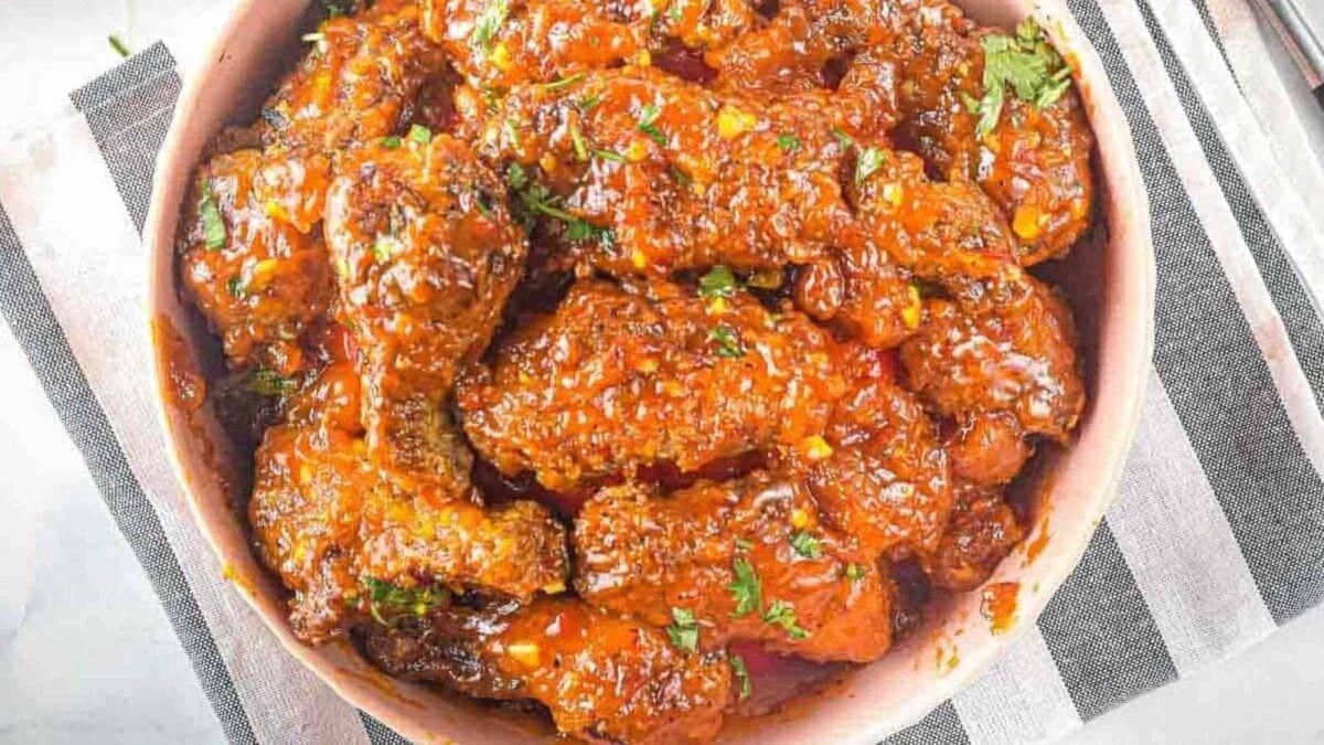 A bowl filled with saucy, glazed chicken wings tossed in chili, garnished with chopped herbs, placed on a striped cloth.