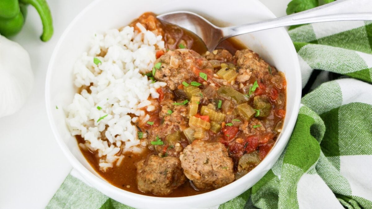 A white bowl holds white rice and meatballs in a tomato-based sauce with vegetables; a spoon rests in the bowl next to a green and white cloth, making it perfect inspiration for new Meatball recipes.