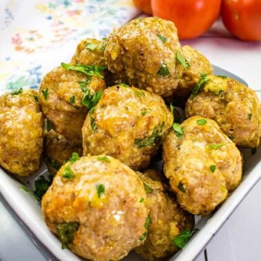 A bowl of cooked meatballs garnished with chopped herbs sits on a white tiled surface, perfect for pairing with spaghetti. Tomatoes and a patterned cloth create a classic Meatball recipes backdrop.
