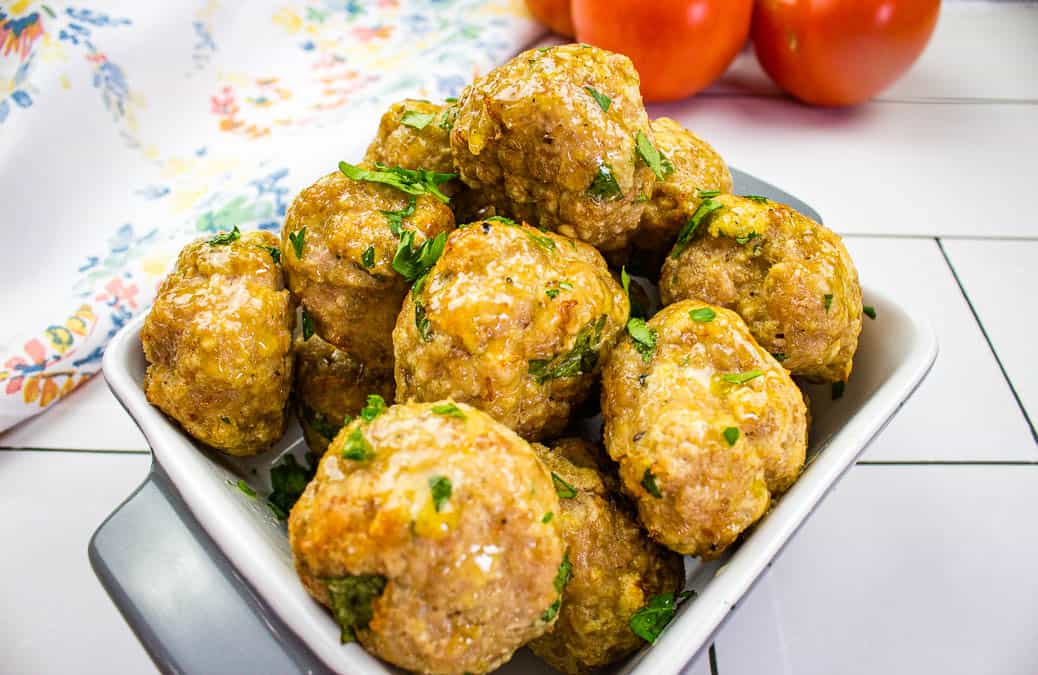 A bowl of cooked meatballs garnished with chopped herbs sits on a white tiled surface, perfect for pairing with spaghetti. Tomatoes and a patterned cloth create a classic Meatball recipes backdrop.