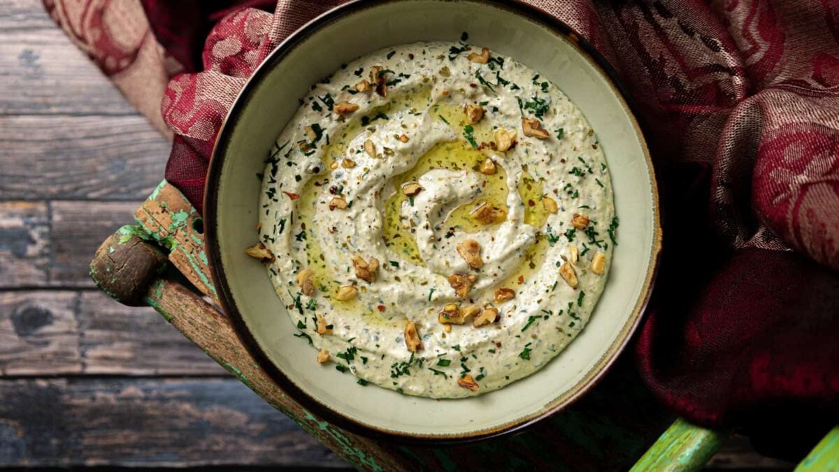 Bowl of baba ganoush garnished with chopped herbs, walnuts, and olive oil, placed on a rustic wooden surface with a red patterned cloth nearby—a delicious addition to your collection of dip recipes and party appetizers.