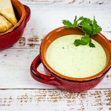 A red ceramic bowl filled with creamy green dip garnished with fresh cilantro, perfect for party appetizers, sits next to a bowl of tortilla chips on a white wooden surface.