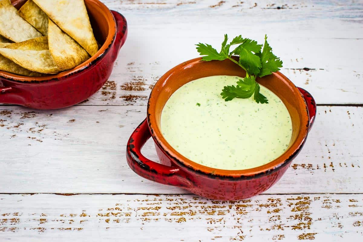 A red ceramic bowl filled with creamy green dip garnished with fresh cilantro, perfect for party appetizers, sits next to a bowl of tortilla chips on a white wooden surface.