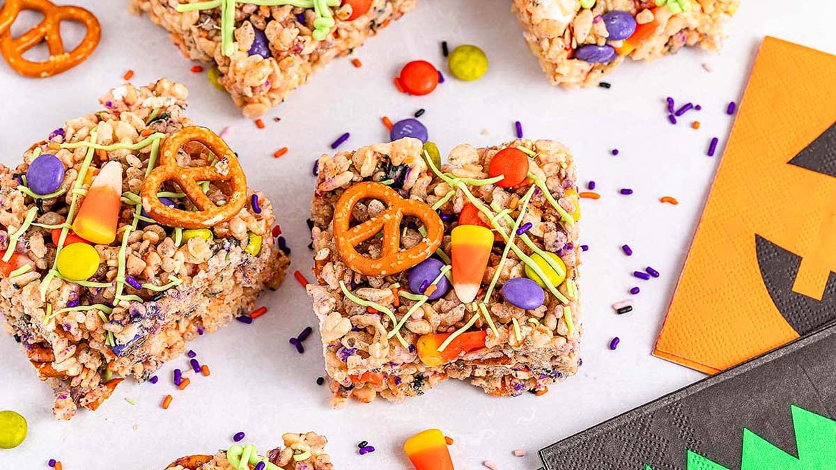 Classic Rice Krispie Treats are topped with candy corn, pretzels, colorful candies, and sprinkles, displayed on a white surface with festive napkins nearby for a spooky seasonal touch.