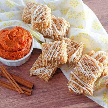 Spooky Rice Krispie Treats drizzled with white icing are arranged on a yellow-patterned cloth next to a bowl of orange dip and two cinnamon sticks on a wooden surface.