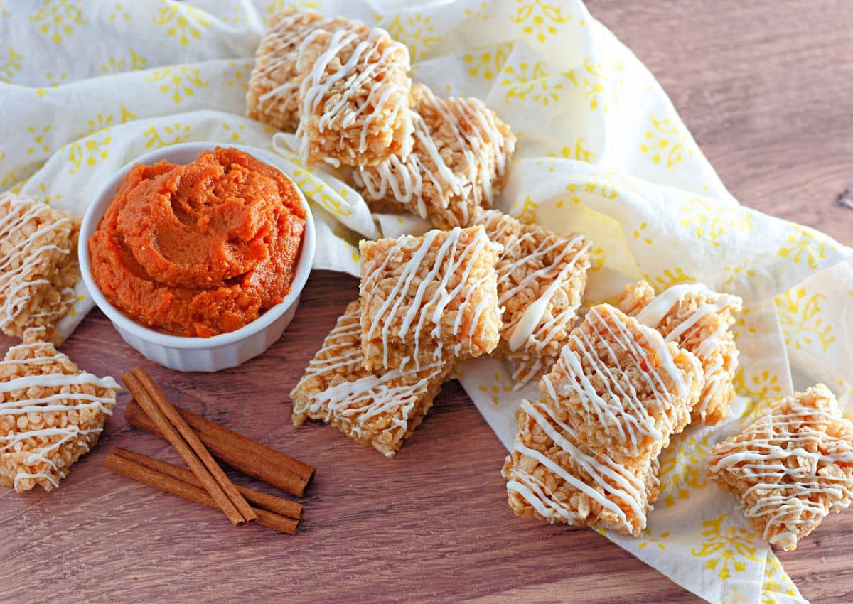 Spooky Rice Krispie Treats drizzled with white icing are arranged on a yellow-patterned cloth next to a bowl of orange dip and two cinnamon sticks on a wooden surface.