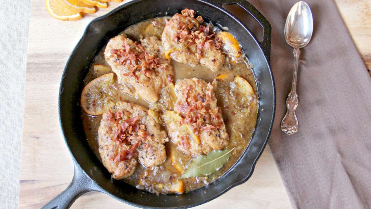 Four seasoned chicken breasts with sauce, garnished with crumbled bacon, orange slices, and a touch of prosciutto, cooked in a cast-iron skillet—a delicious nod to sophisticated prosciutto recipes. A spoon and napkin are beside the skillet.