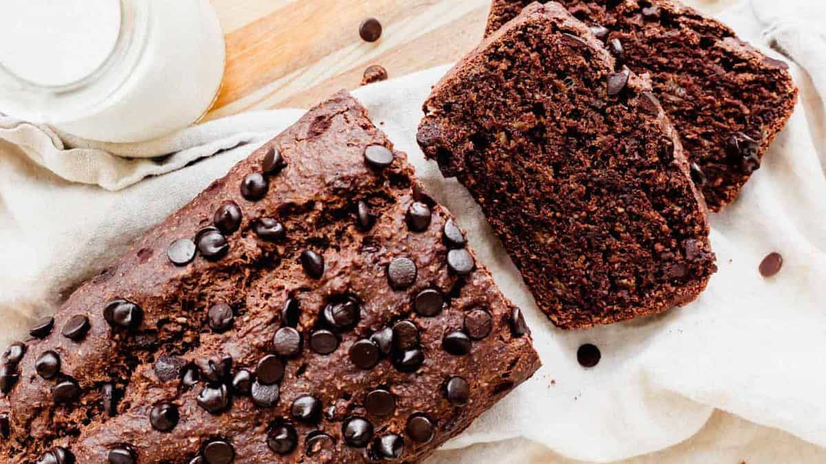 A loaf of chocolate chip bread on a cloth, with two slices cut and placed beside it, and a glass of milk in the background—perfect for anyone looking for delicious Gluten Free Baking.