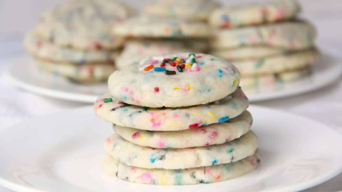 A stack of homemade round sugar cookies with colorful sprinkles is placed on a white plate, with more cookies stacked on plates in the background.