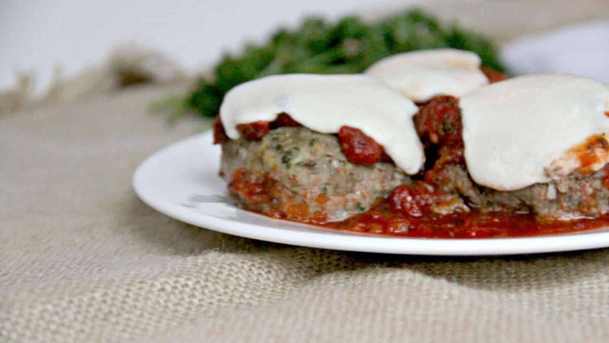 Three meatballs topped with melted mozzarella cheese and tomato sauce on a white plate, perfect for everyday meals, with a burlap tablecloth and herbs blurred in the background.