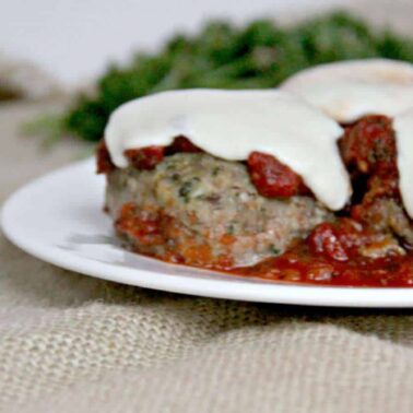 Three meatballs topped with melted mozzarella cheese and tomato sauce on a white plate, perfect for everyday meals, with a burlap tablecloth and herbs blurred in the background.