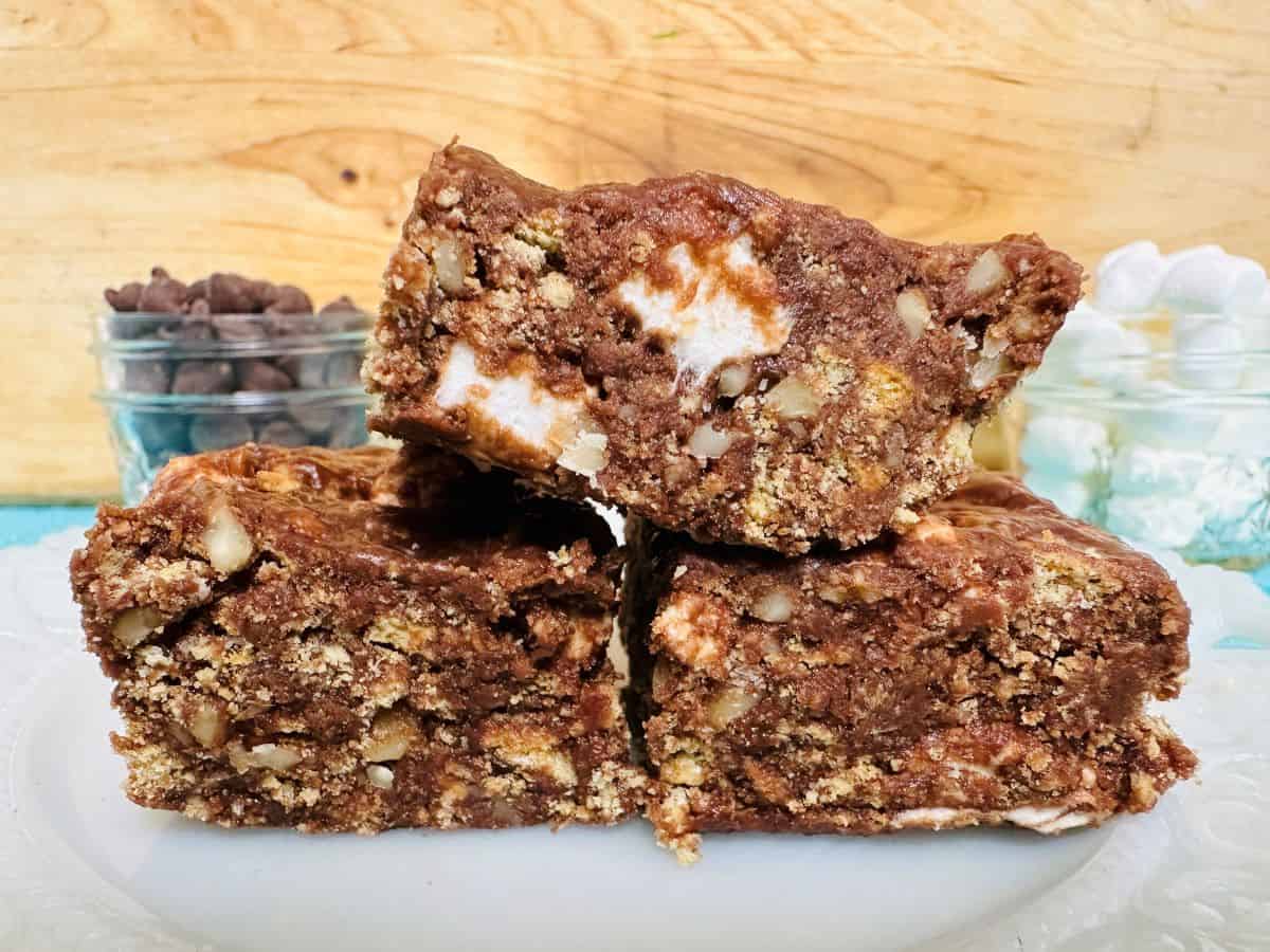 Three rocky road dessert bars with visible nuts and marshmallows are stacked on a white plate; chocolate chips and marshmallows sit in the background—perfect for fans of egg-free recipes.