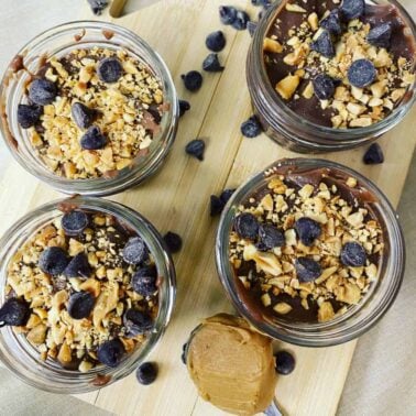 Four jars of chocolate pudding topped with chopped nuts and chocolate chips, arranged on a wooden board with a spoonful of peanut butter—one of our most satisfying, egg-free recipes for a rich and indulgent treat.