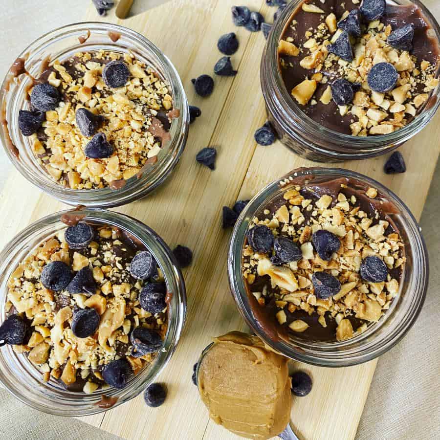 Four jars of chocolate pudding topped with chopped nuts and chocolate chips, arranged on a wooden board with a spoonful of peanut butter—one of our most satisfying, egg-free recipes for a rich and indulgent treat.