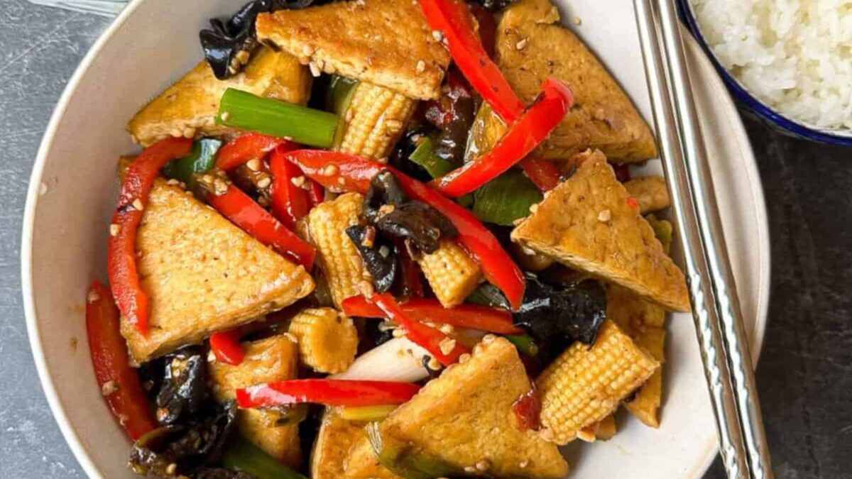 A bowl of stir-fried tofu with red bell peppers, baby corn, mushrooms, and green onions, served with chopsticks and a side of white rice—perfect for those seeking delicious gluten-free meals.