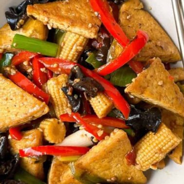 A bowl of stir-fried tofu with red bell peppers, baby corn, mushrooms, and green onions, served with chopsticks and a side of white rice—perfect for those seeking delicious gluten-free meals.
