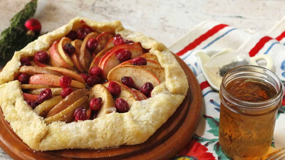 A rustic fruit galette with apple slices and cranberries on a wooden board, next to a jar of honey on a colorful cloth—one of those easy treats that transforms simple ingredients into a charming dessert.