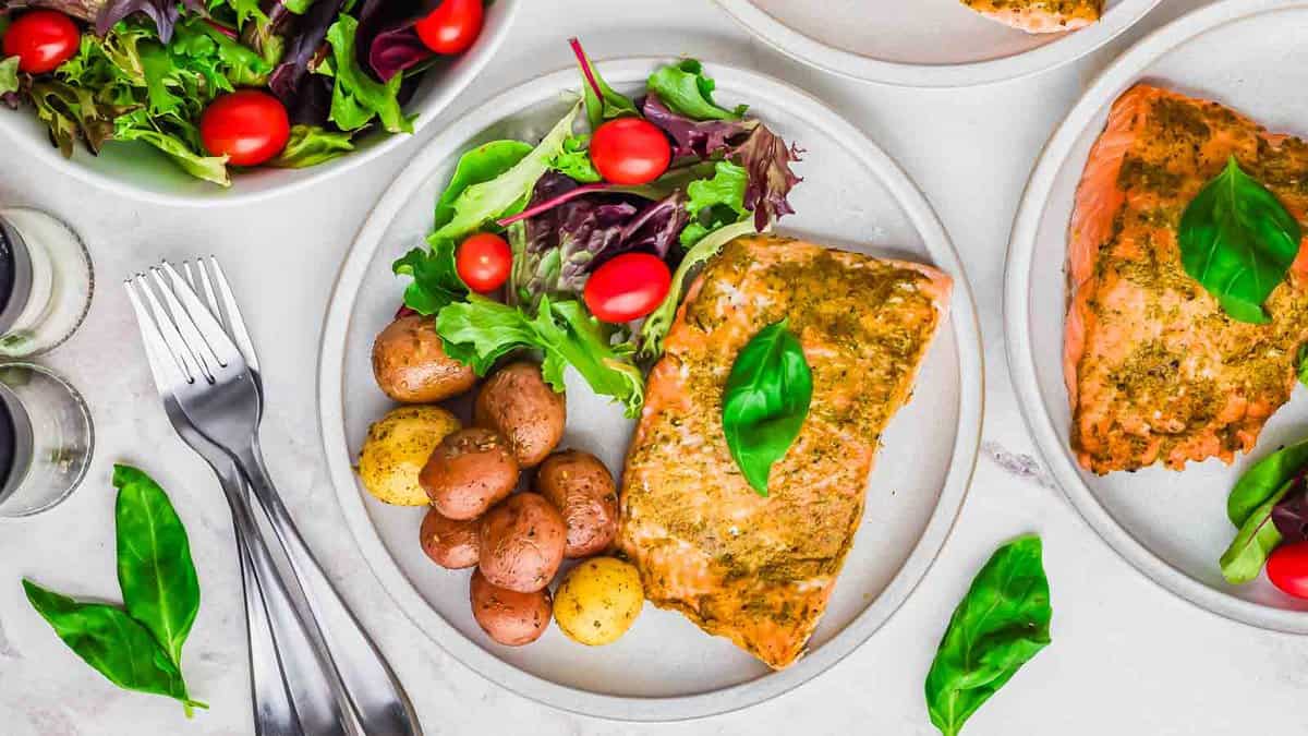 A plate with baked salmon, mixed salad with cherry tomatoes, and roasted baby potatoes, garnished with basil leaves—one of our favorite filling low carb meals. Forks and extra greens are nearby.