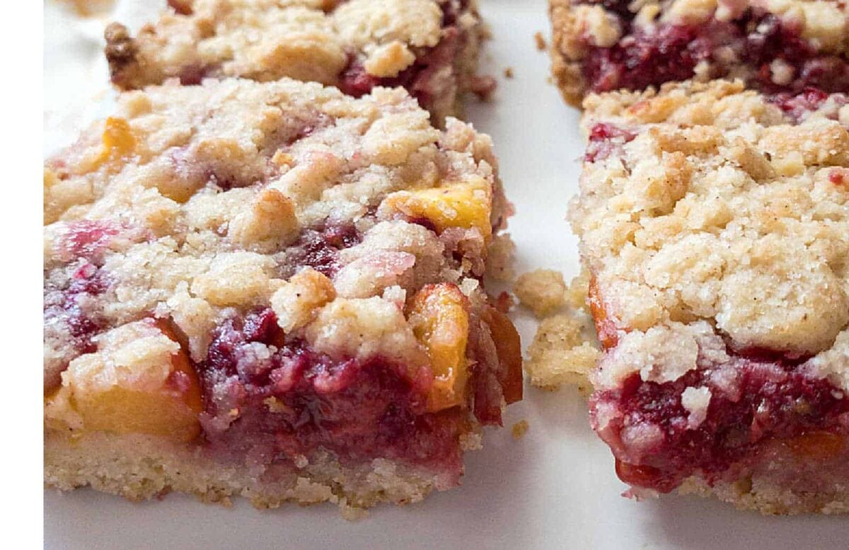 Close-up of fruit crumble bars with a crumbly topping and a visible layer of mixed fruit filling, including peaches and berries, on a white surface—easy treats that look like fancy desserts with zero effort.