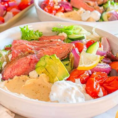 A bowl filled with sliced steak, avocado, lettuce, tomatoes, cucumber, red onion, feta cheese, olives, hummus, and tzatziki sauce—perfect for enjoying a fresh twist on classic steakhouse dinners.