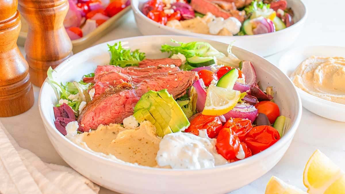 A bowl filled with sliced steak, avocado, lettuce, tomatoes, cucumber, red onion, feta cheese, olives, hummus, and tzatziki sauce—perfect for enjoying a fresh twist on classic steakhouse dinners.