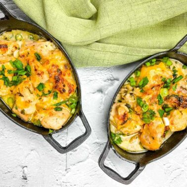 Two oval cast iron dishes filled with creamy baked chicken and vegetables, topped with melted cheese and fresh herbs—these chicken casseroles make perfect family meals. Placed on a white surface next to a green cloth.
