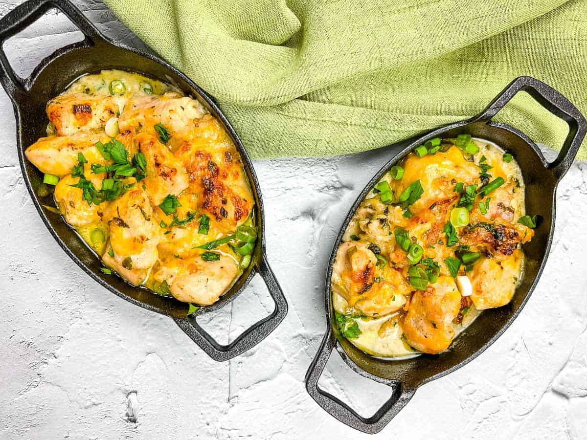 Two oval cast iron dishes filled with creamy baked chicken and vegetables, topped with melted cheese and fresh herbs—these chicken casseroles make perfect family meals. Placed on a white surface next to a green cloth.