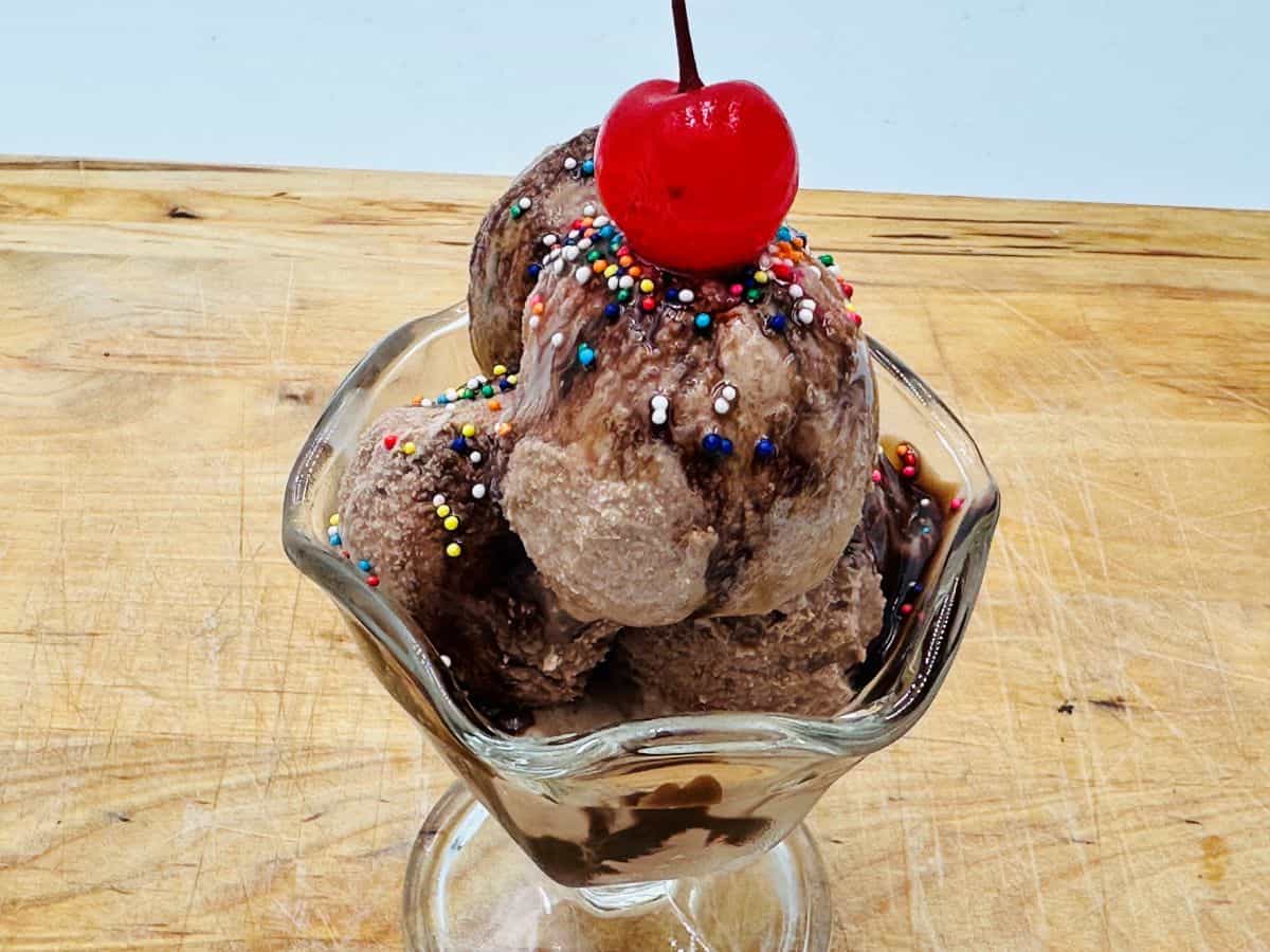 Three scoops of chocolate ice cream in a glass dish, topped with chocolate syrup, colorful sprinkles, and a maraschino cherry on a wooden surface—a perfect choice for lovers of sunshine desserts.