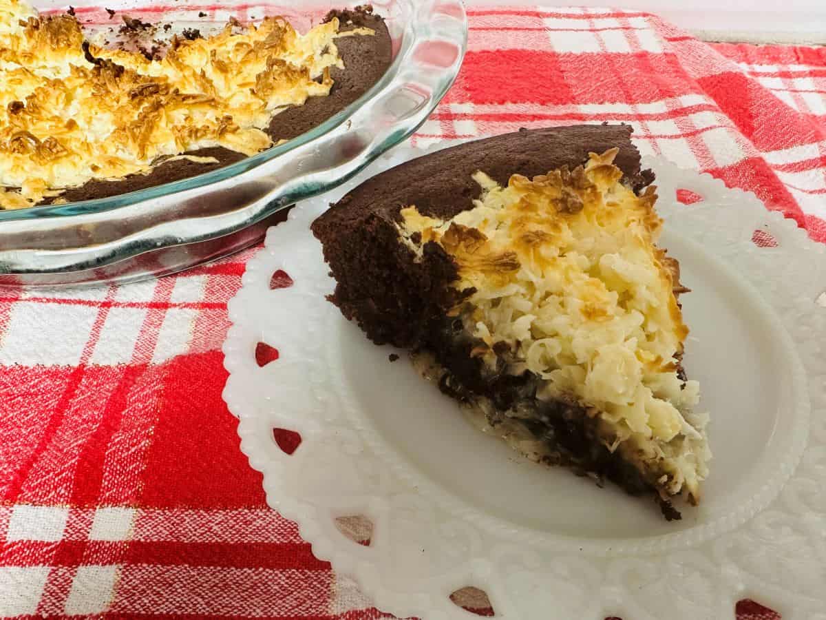 A slice of chocolate and coconut pie on a white plate, with the rest of the pie in a glass dish in the background, set on a red plaid tablecloth—perfect for sunshine desserts and sweet summer gatherings.