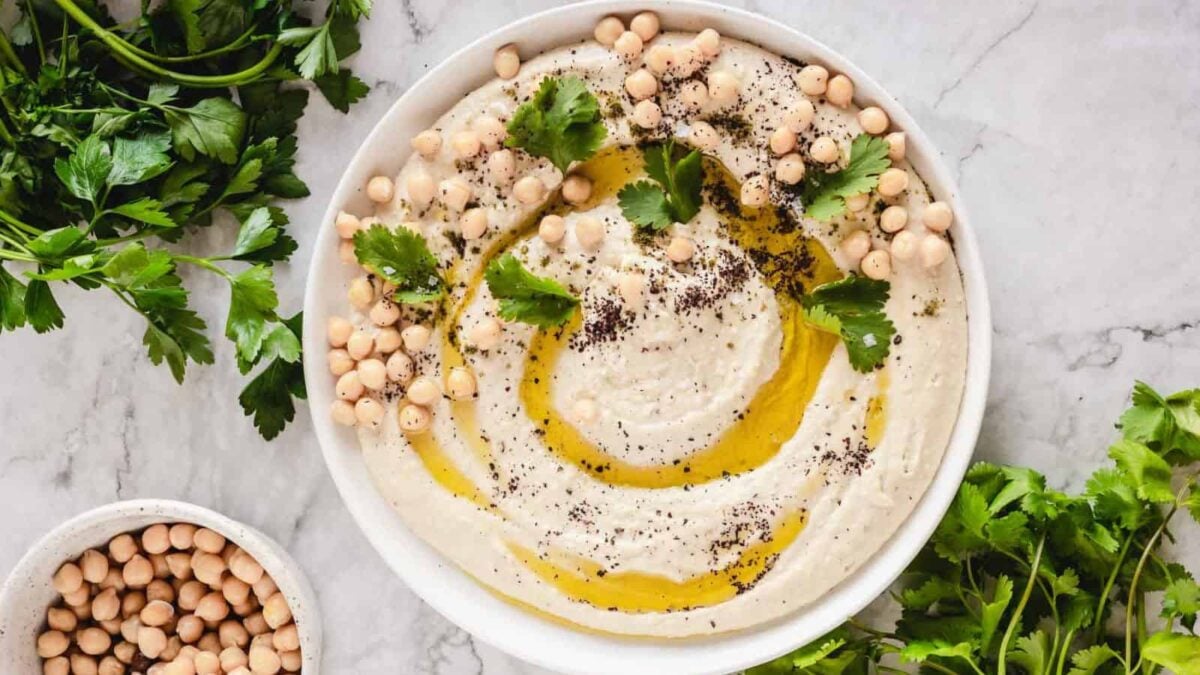 A bowl of hummus garnished with chickpeas, olive oil, herbs, and spices, surrounded by fresh parsley and a small bowl of chickpeas on a marble surface—a delicious plant-based option for vegan recipes and meat-eaters alike.