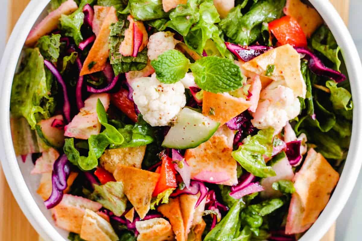 A bowl of vegan fattoush salad with chopped lettuce, red cabbage, cucumber, tomato, radish, cauliflower, pita chips, and fresh mint leaves—a fresh option that even meat-eaters will love.