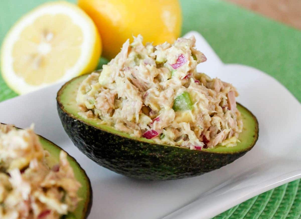 Half an avocado filled with tuna salad, garnished with diced vegetables, is the perfect addition to your lineup of refreshing seafood recipes—served on a white plate with a sliced lemon in the background.