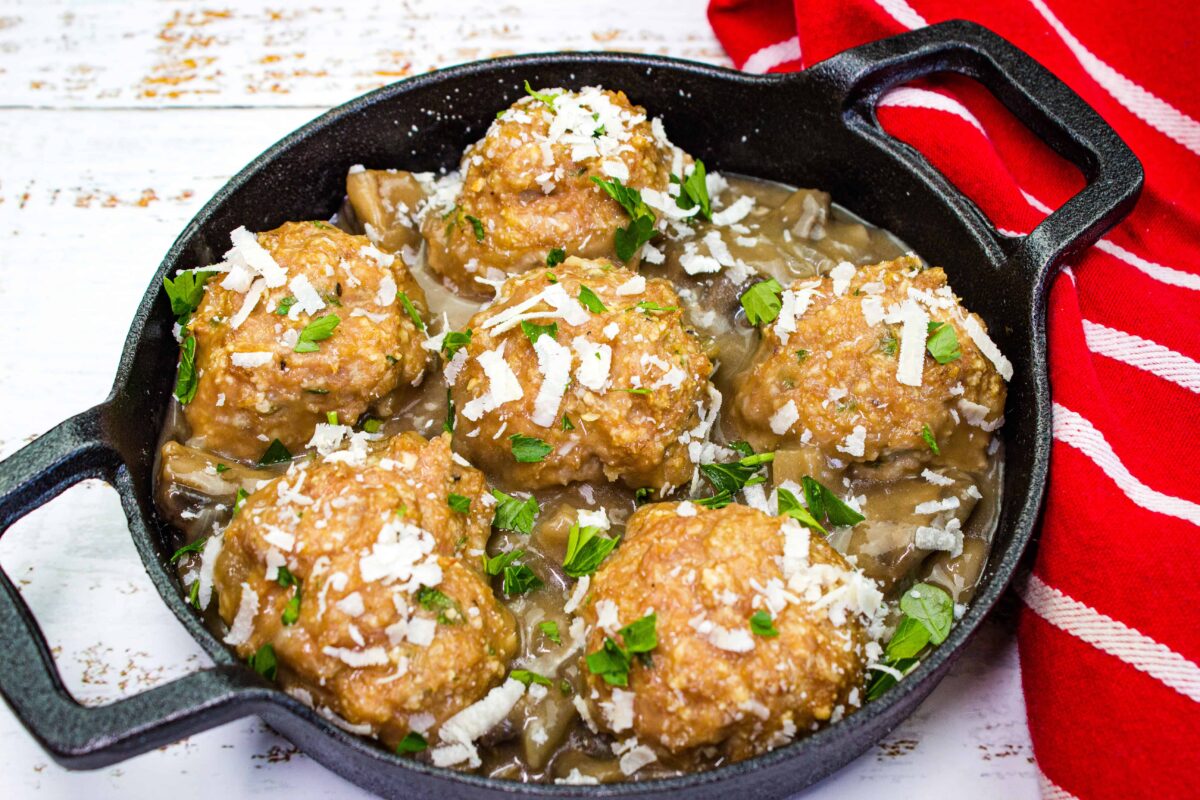 Six large meatballs in sauce topped with grated cheese and chopped parsley in a black cast iron skillet, perfect for those seeking filling low carb dinners, served next to a red and white striped cloth.