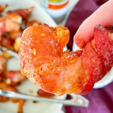 A hand holding a piece of crispy, cooked bacon shaped like a heart, with more bacon pieces and utensils on a plate in the background—perfect inspiration for vacation recipes or creative breakfast ideas.