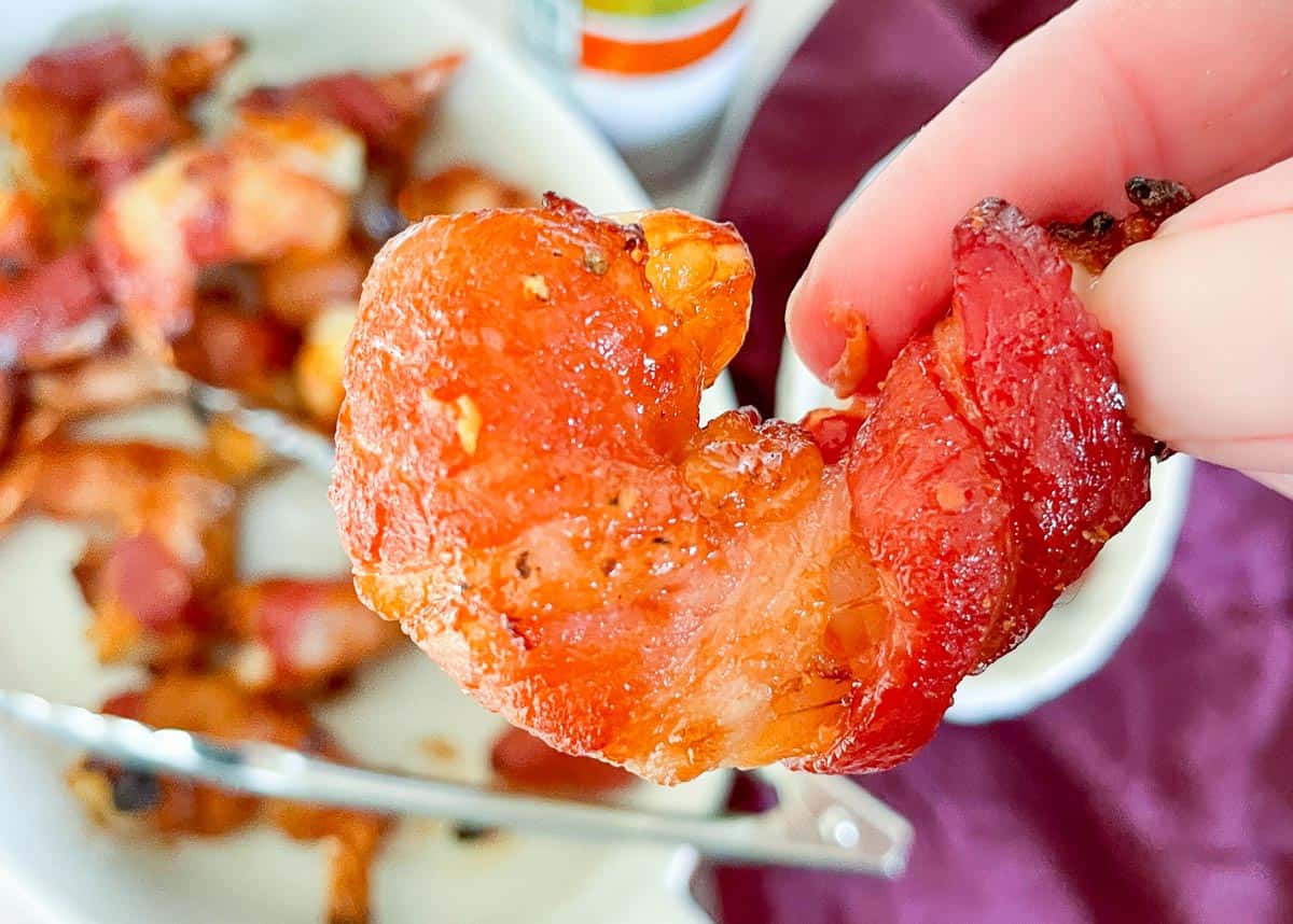 A hand holding a piece of crispy, cooked bacon shaped like a heart, with more bacon pieces and utensils on a plate in the background—perfect inspiration for vacation recipes or creative breakfast ideas.