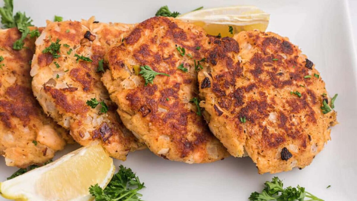 Four golden-brown salmon patties garnished with chopped parsley, served with fresh lemon wedges on a white plate—a delicious nod to vintage main dishes.