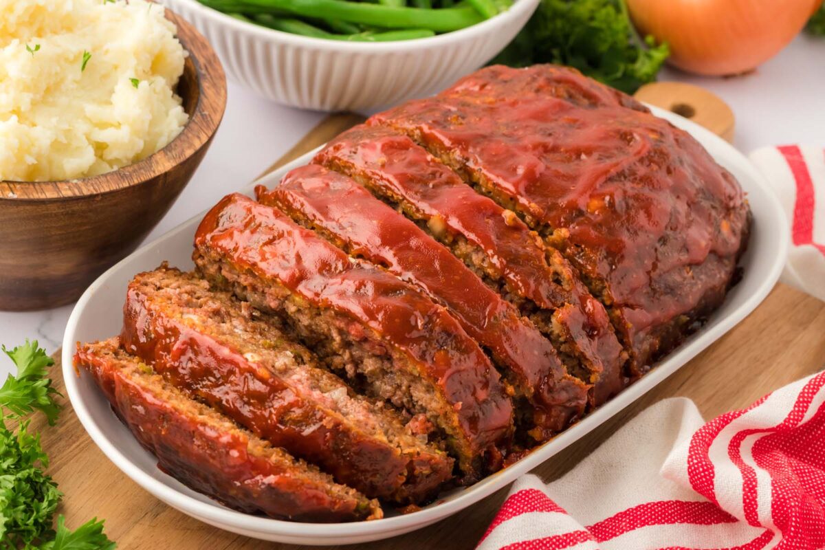 A sliced meatloaf with ketchup glaze—a staple of classic recipes and retro dinners—is served on a white dish, accompanied by bowls of mashed potatoes and green beans in the background.