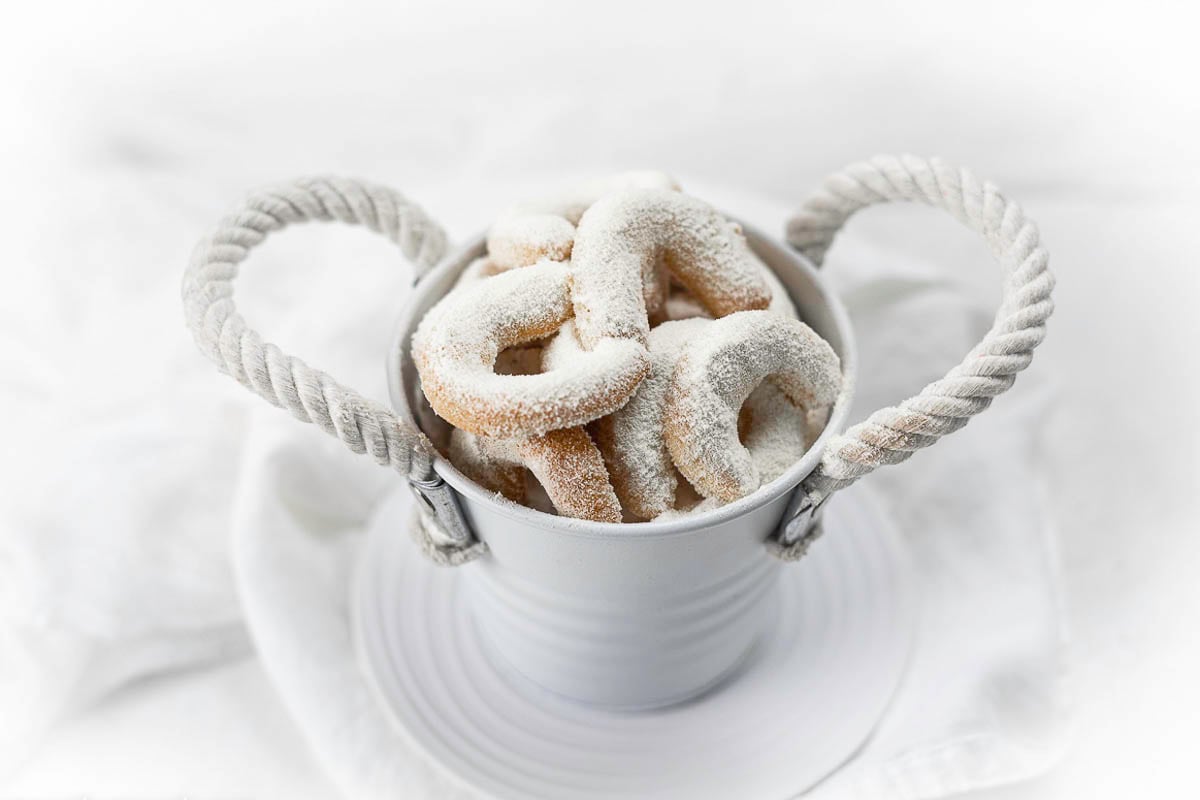 A small white bucket filled with powdered sugar-coated, egg free cookies in crescent shapes, placed on a white plate and white fabric background.