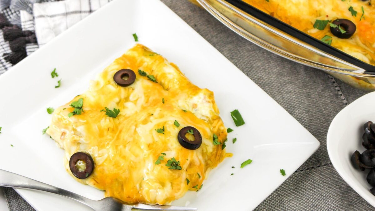 A serving of cheesy enchiladas garnished with sliced black olives and chopped herbs on a white plate, perfect for those who love effortless meals and baked chicken dinners, with a fork placed beside it.
