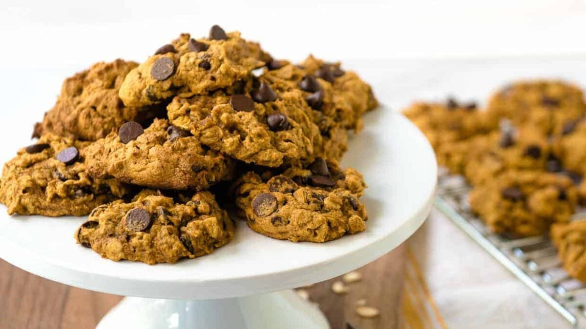 A pile of chewy, eggless cookies sits on a white cake stand, with more cookies cooling on a rack in the background.