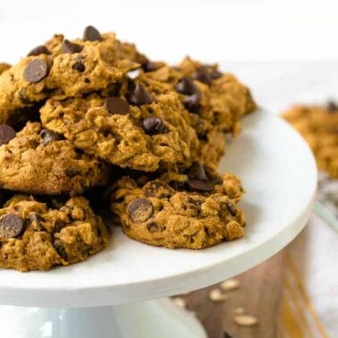 A pile of chewy, eggless cookies sits on a white cake stand, with more cookies cooling on a rack in the background.