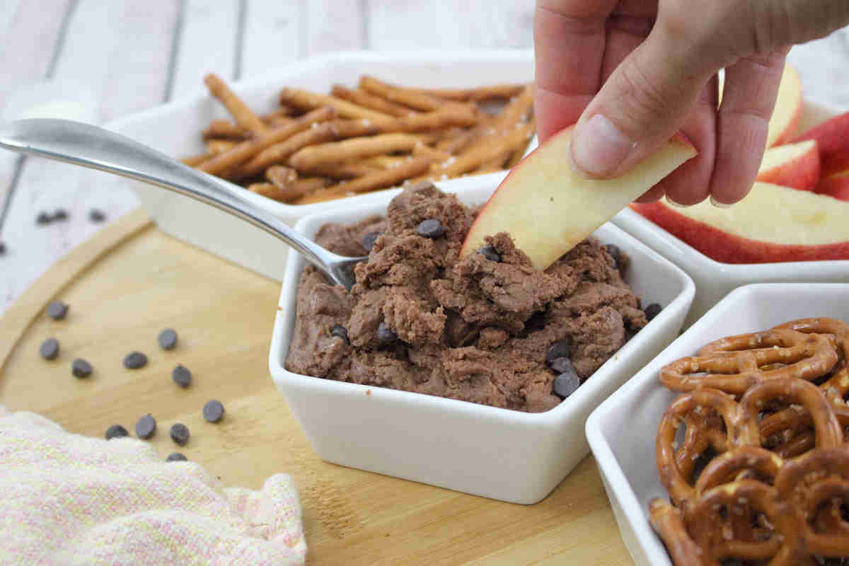 A hand dips an apple slice into a bowl of chocolate chip dessert dip, surrounded by pretzels and more apple slices on a wooden board—perfect inspiration for lovers of decadent desserts.