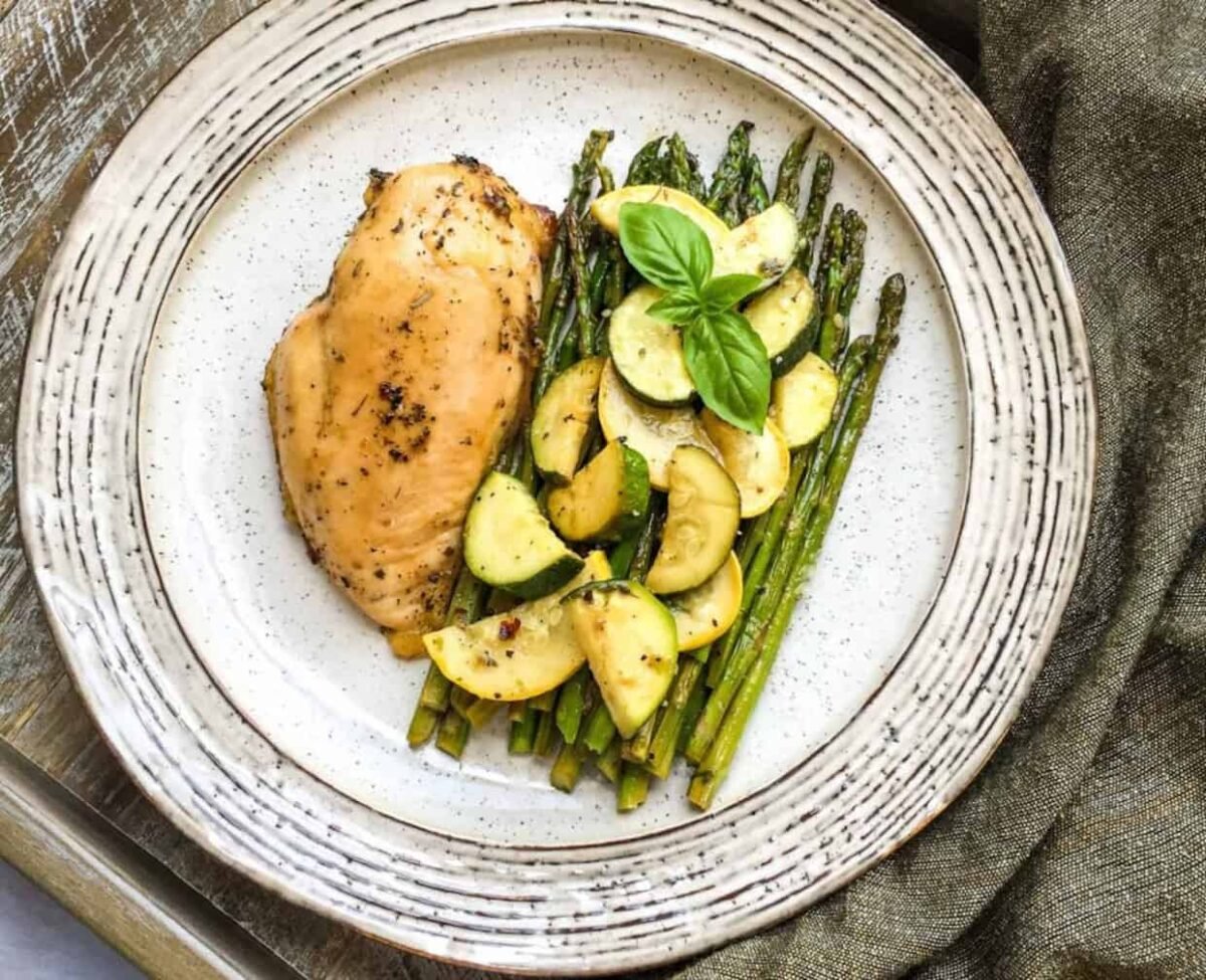 Grilled chicken breast served with asparagus and sautéed zucchini, garnished with fresh basil, on a round ceramic plate—perfect for fans of easy dinners and delicious one-pan recipes.