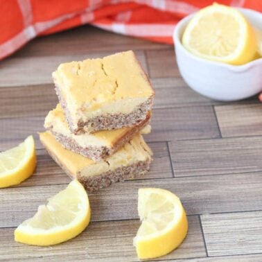 Three lemon bars are stacked on a wooden surface with lemon wedges nearby, a small bowl of lemon slices, and an orange plaid cloth in the background—an inviting display of indulgent desserts.