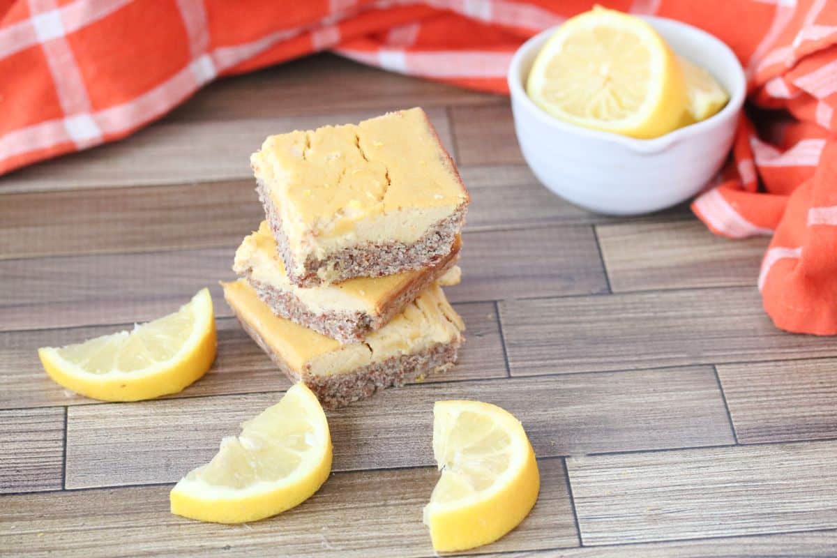 Three lemon bars are stacked on a wooden surface with lemon wedges nearby, a small bowl of lemon slices, and an orange plaid cloth in the background—an inviting display of indulgent desserts.