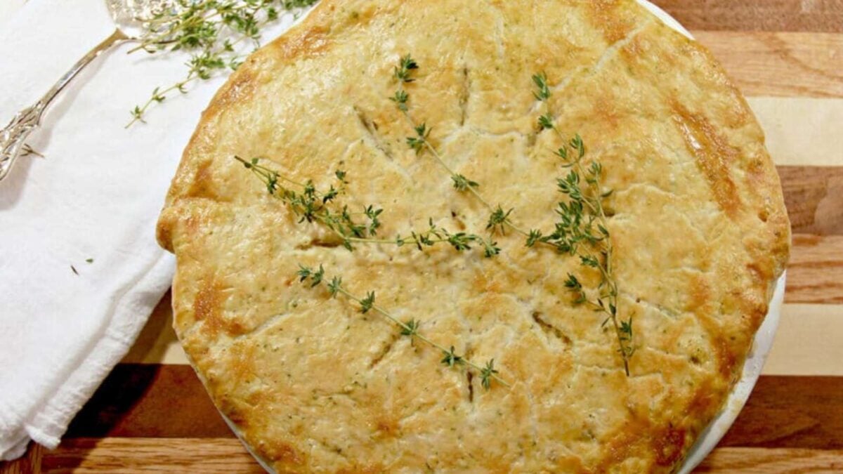 A golden-brown savory pie topped with sprigs of fresh thyme, placed on a wooden surface next to a white napkin and a serving utensil—a delicious showcase of classic pie fillings.