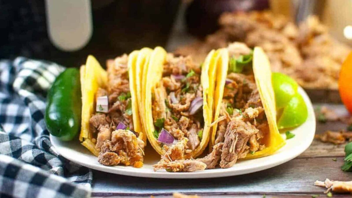 A plate of three tacos filled with shredded meat, chopped onions, and cilantro, garnished with a green chili and lime wedges, makes for easy dinners on a wooden table.