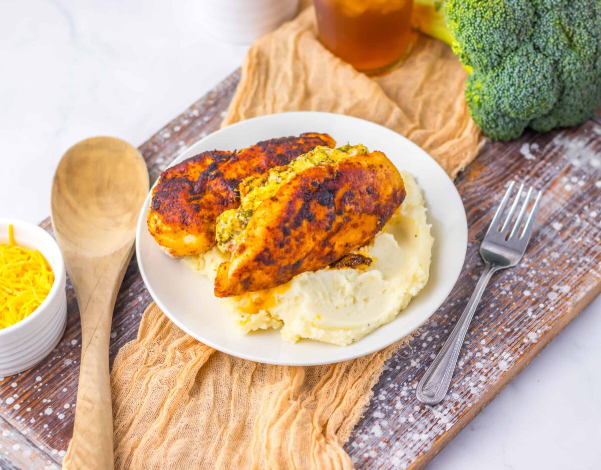 A plate with two stuffed, grilled chicken breasts on mashed potatoes sits next to a fork, wooden spoon, shredded cheese, broccoli, and a glass of iced tea—perfect inspiration for tasty dinners and flavorful meals.