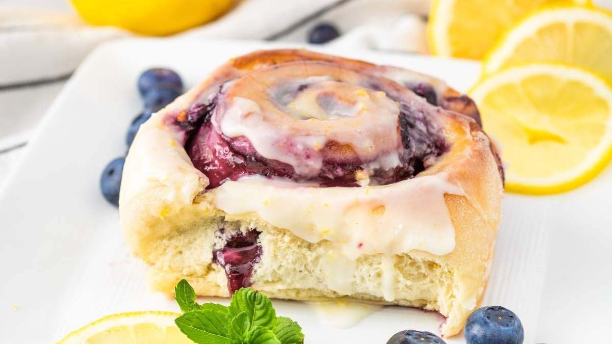A blueberry sweet roll topped with white icing, garnished with lemon slices, blueberries, and a mint sprig on a white plate—perfect for easy breakfast recipes or relaxing weekend breakfasts.