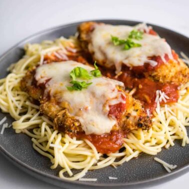 A gray plate with two pieces of chicken parmesan topped with marinara sauce and melted cheese, served over a bed of spaghetti and garnished with basil—one of those air fryer recipes sure to delight your taste buds.